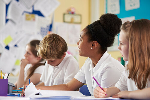 Four pupils sat at a table in school, looking at their books or up at out of shot.